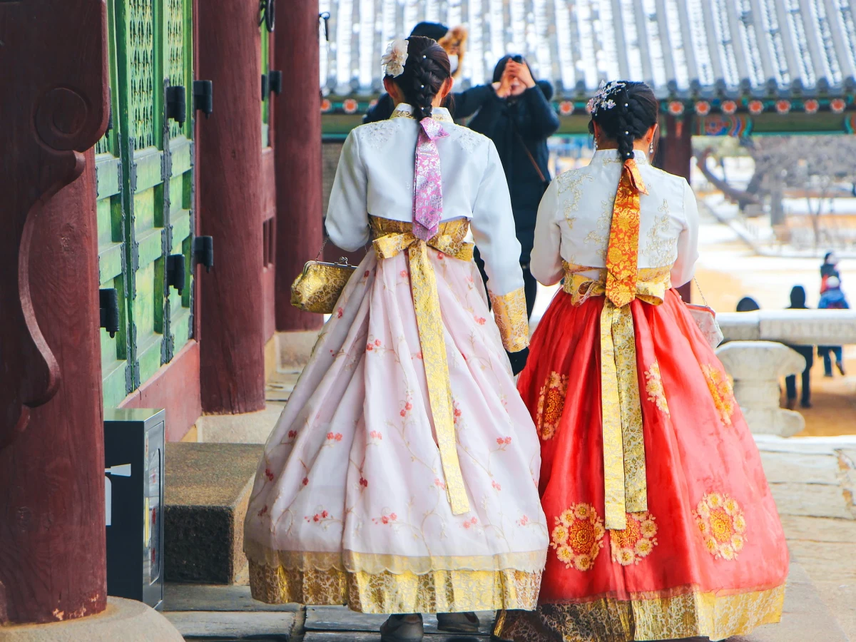Zwei Personen in traditionellen koreanischen Hanbok-Kleidern stehen vor einem historischen Gebäude, eine in einem pastellfarbenen Kleid mit gelber Schärpe und eine in einem roten Kleid mit goldener Verzierung, vor einer traditionellen koreanischen Architektur.