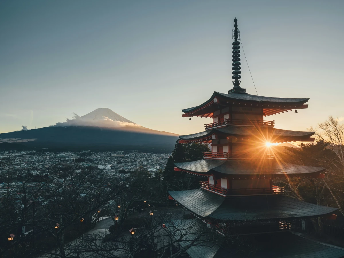 Chureito-Pagode mit Blick auf den schneebedeckten Fuji-Berg bei Sonnenaufgang. Mehrere Stockwerke des roten Tempels im Vordergrund, mit Sonnenstern, der zwischen den Dachebenen hindurchscheint. Weite Stadtlandschaft und Bäume im Hintergrund unter einem klaren Morgenhimmel.