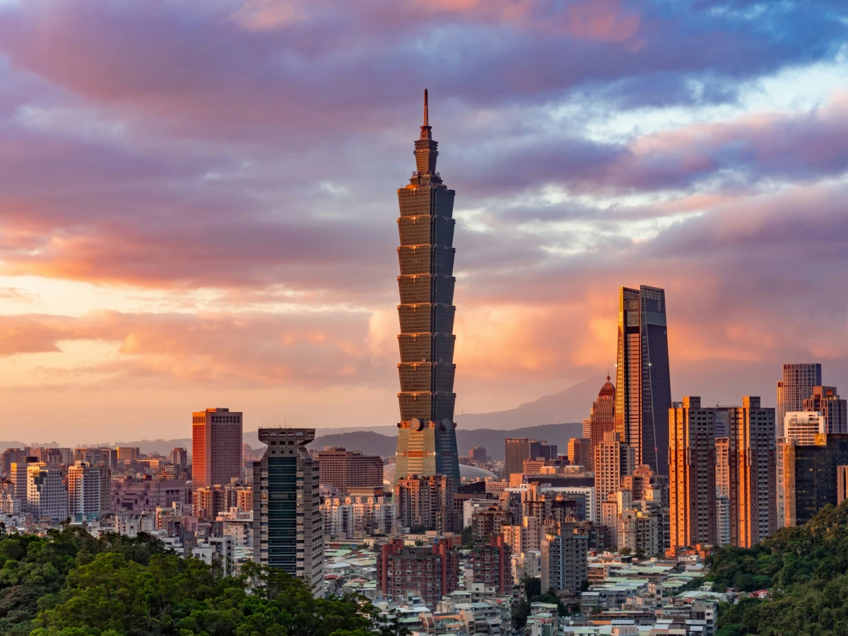 Skyline von Taipeh bei Sonnenuntergang mit dem markanten Taipei 101 Wolkenkratzer im Vordergrund, umgeben von modernen Hochhäusern und einem dramatischen Himmel in Pastellfarben, im Hintergrund bewaldete Berge.