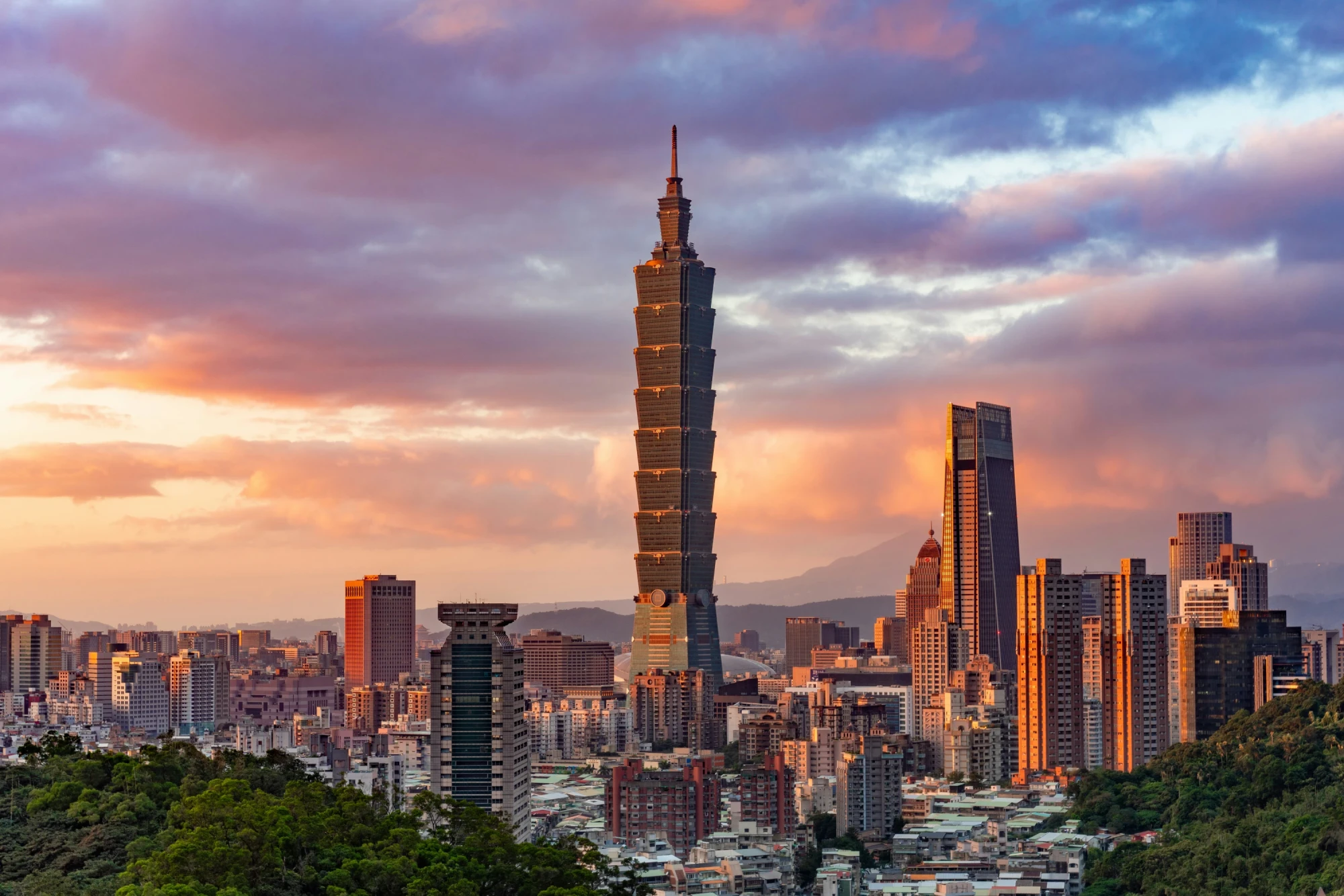 Skyline von Taipeh bei Sonnenuntergang mit dem markanten Taipei 101 Wolkenkratzer im Vordergrund, umgeben von modernen Hochh&auml;usern und einem dramatischen Himmel in Pastellfarben, im Hintergrund bewaldete Berge.