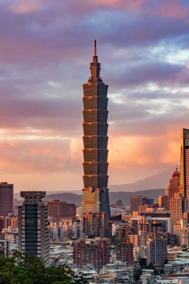 Skyline von Taipeh bei Sonnenuntergang mit dem markanten Taipei 101 Wolkenkratzer im Vordergrund, umgeben von modernen Hochhäusern und einem dramatischen Himmel in Pastellfarben, im Hintergrund bewaldete Berge.