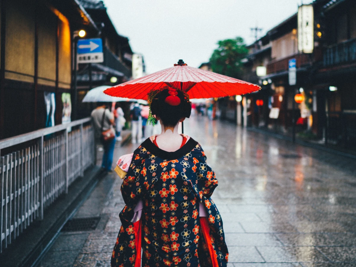 Rückansicht einer Person in traditioneller japanischer Kleidung mit einem roten Schirm, die an einem regnerischen Tag eine alte Straße in Kyoto entlang geht, umgeben von historischen Holzgebäuden und gedämpften Lichtern.