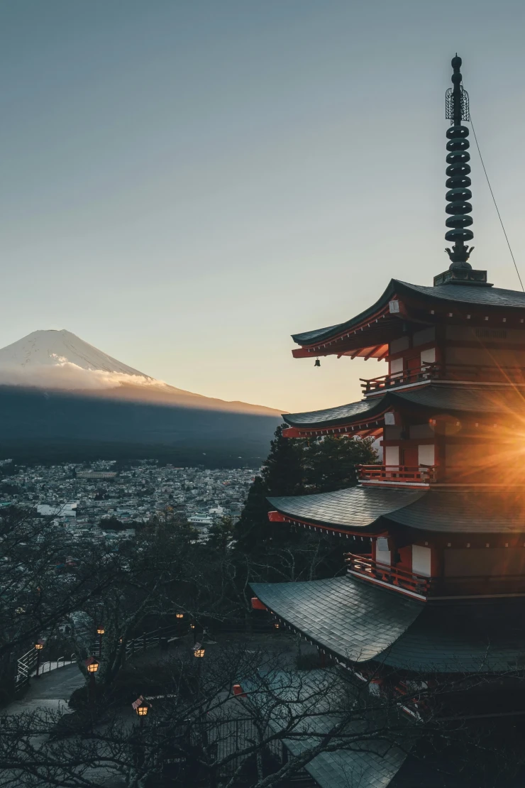 Chureito-Pagode mit Blick auf den schneebedeckten Fuji-Berg bei Sonnenaufgang. Mehrere Stockwerke des roten Tempels im Vordergrund, mit Sonnenstern, der zwischen den Dachebenen hindurchscheint. Weite Stadtlandschaft und Bäume im Hintergrund unter einem klaren Morgenhimmel.