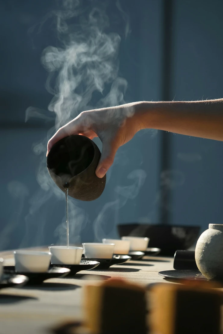 Dampfende Tasse wird von einer Hand über mehrere Teetassen in einer stimmungsvollen, bläulichen Umgebung gegossen. Zartes Dampfwölkchen steigt auf, während heißes Getränk eingeschenkt wird.
