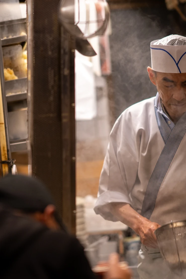 Weißgekleideter Koch in einer traditionellen japanischen Ramenküche bereitet sorgfältig Nudeln zu, umgeben von Küchengeräten und gedämpfter Beleuchtung. Die Atmosphäre ist geschäftig und authentisch, mit Blick auf die Zubereitung der Speisen.