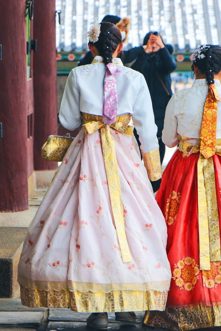 Zwei Personen in traditionellen koreanischen Hanbok-Kleidern stehen vor einem historischen Gebäude, eine in einem pastellfarbenen Kleid mit gelber Schärpe und eine in einem roten Kleid mit goldener Verzierung, vor einer traditionellen koreanischen Architektur.
