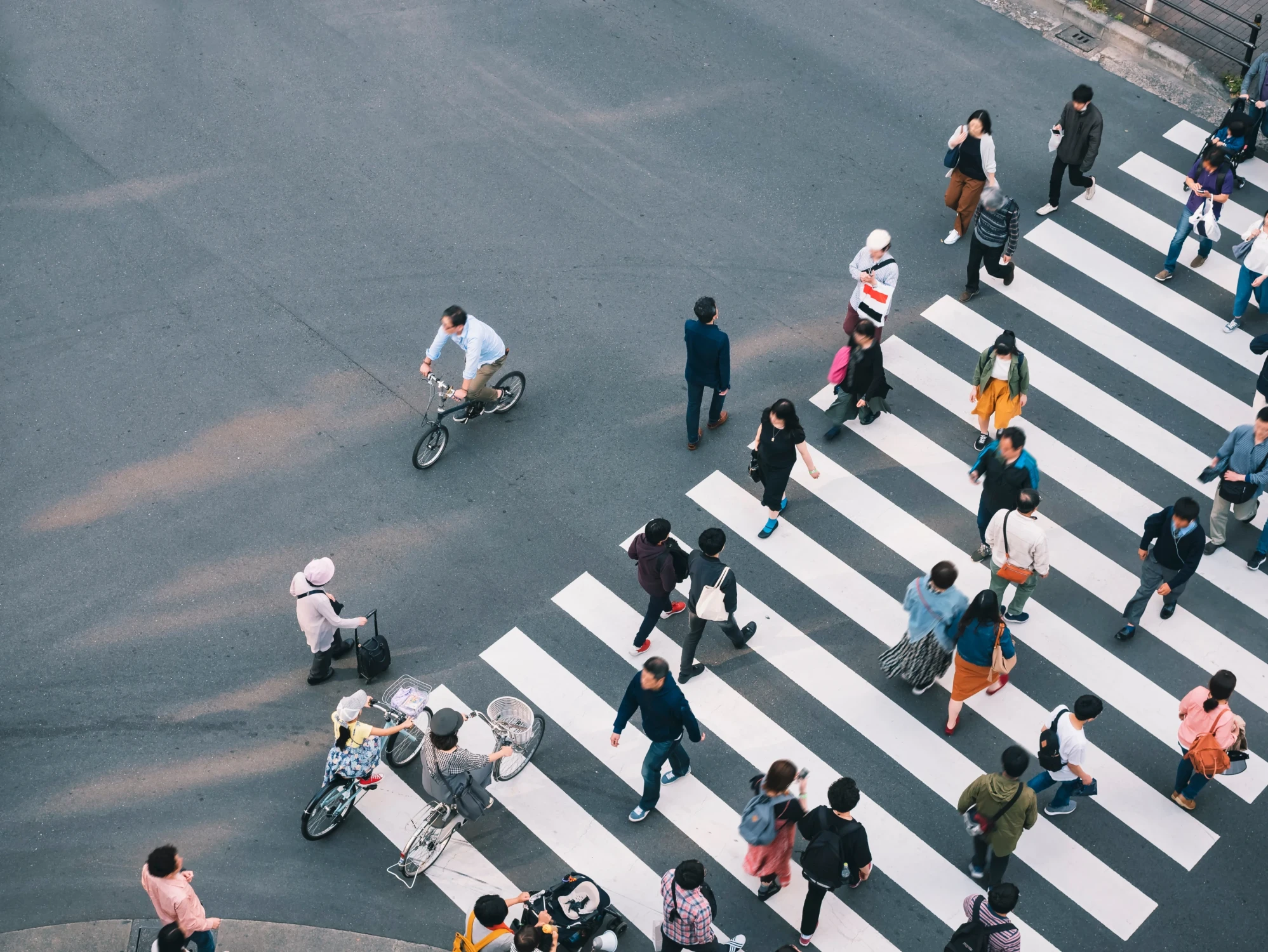 Luftaufnahme einer belebten Fußgängerüberquerung in einer Stadt mit Menschen in verschiedenen Kleidungsfarben, die einen Zebrastreifen überqueren, während ein Radfahrer im Hintergrund fährt.
