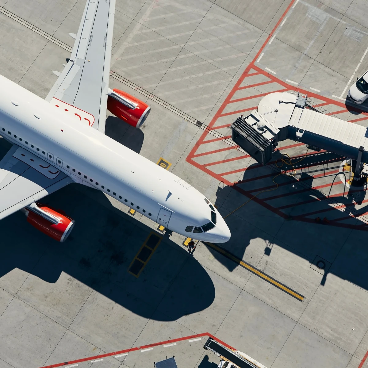 Luftaufnahme eines weißen Passagierflugzeugs an einem Flughafengate mit Flugzeugbrücke. Rote Triebwerke und Seitenleitwerk sind sichtbar, während Bodenpersonal und Fahrzeuge um das Flugzeug herum positioniert sind.