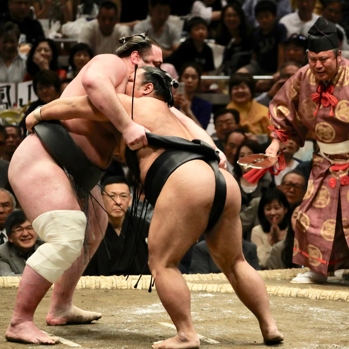Zwei Sumō-Ringer kämpfen intensiv auf einem Wettkampfring, umgeben von aufgeregten Zuschauern und einem traditionell gekleideten Schiedsrichter. Der Kampf findet in einer vollen Halle statt, mit Spannung und Konzentration in der Luft.