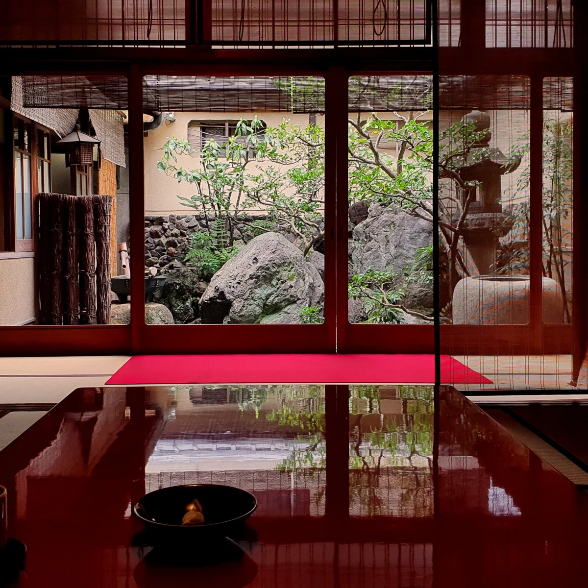 Traditioneller japanischer Raum mit glänzend rotem Boden, Schiebetüren und Blick auf einen zen-artigen Steingarten mit Baum und Steinlaterne, der Ruhe und Eleganz ausstrahlt.