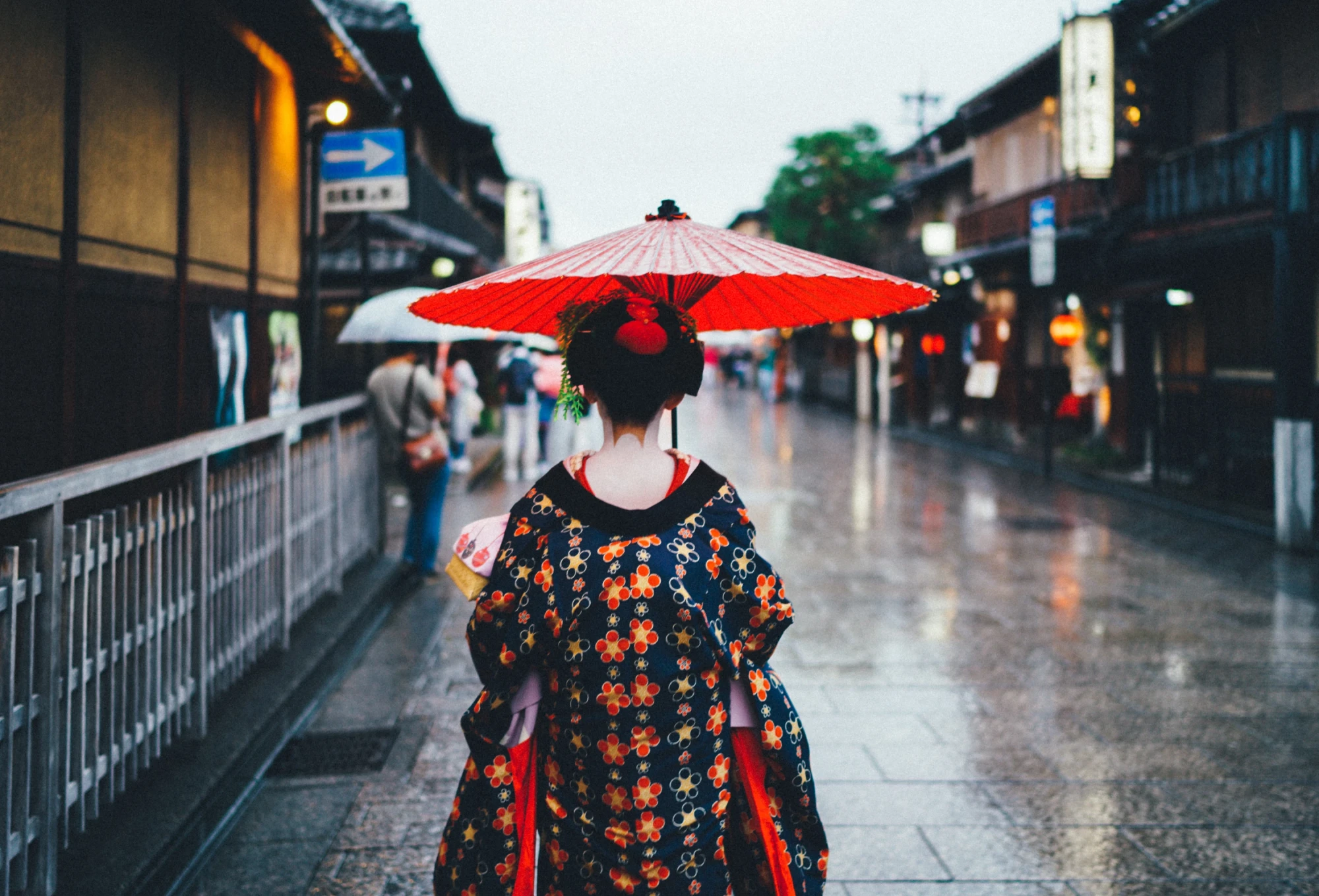 Rückansicht einer Person in traditioneller japanischer Kleidung mit einem roten Schirm, die an einem regnerischen Tag eine alte Straße in Kyoto entlang geht, umgeben von historischen Holzgebäuden und gedämpften Lichtern.
