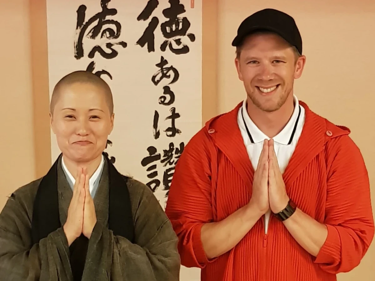 Eine Frau in traditioneller Zen-Kleidung und ein lächelnder Mann in roter Jacke stehen nebeneinander und halten respektvoll die Hände in Gebetshaltung vor einer Kalligraphie-Tapete.