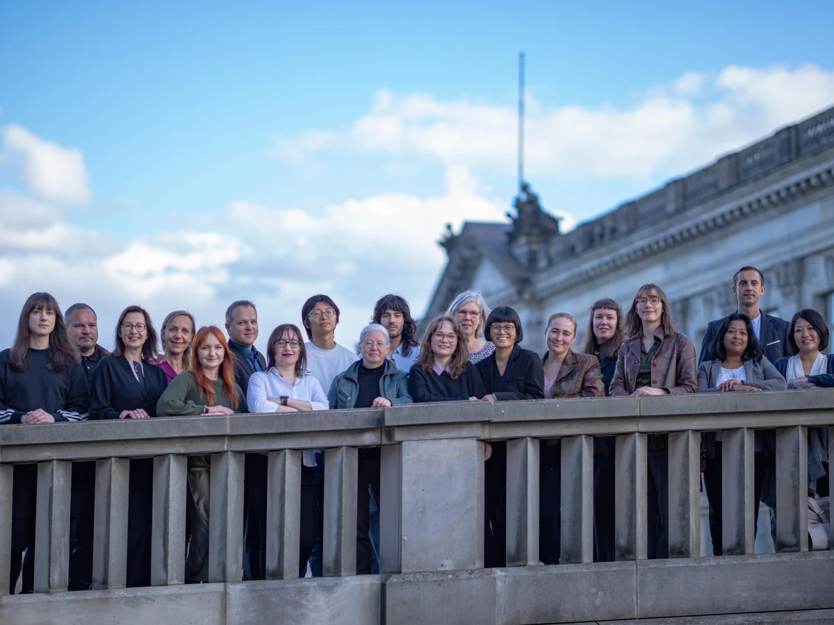 Gruppenaufnahme des J-Navigator Teams vor einer künstlerischen Wandmalerei, bestehend aus etwa 15 Personen unterschiedlichen Alters und Hintergrundes, gekleidet in verschiedene Casual- und Business-Casual-Outfits, die freundlich und professionell in die Kamera blicken.