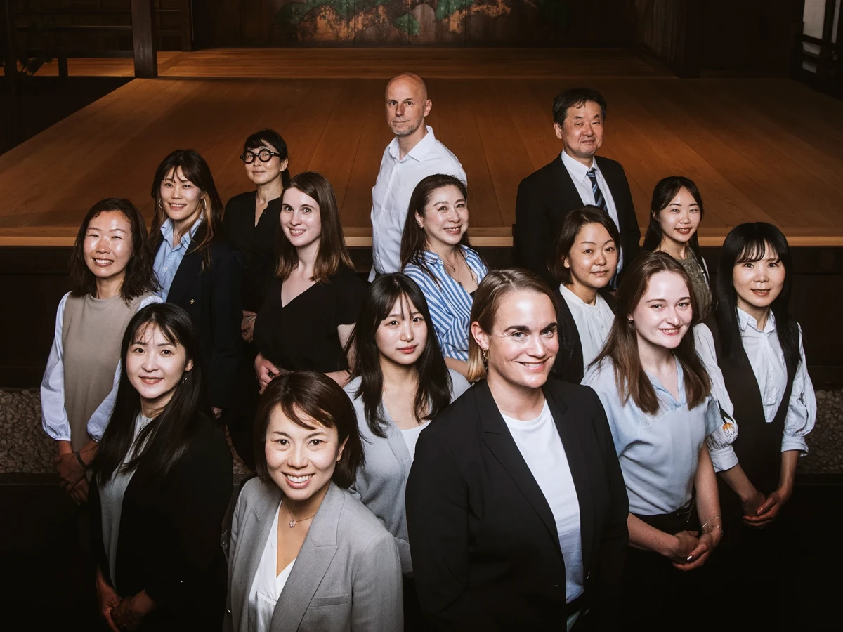 Professionelles Gruppenporträt eines internationalen Teams mit 13 Mitarbeitenden, das in zwei Reihen posiert und eine Mischung aus Männern und Frauen unterschiedlicher Herkunft zeigt, gekleidet in geschäftliche Berufskleidung in gedeckten Farbtönen vor einem warmen, holzvertäfelten Hintergrund.